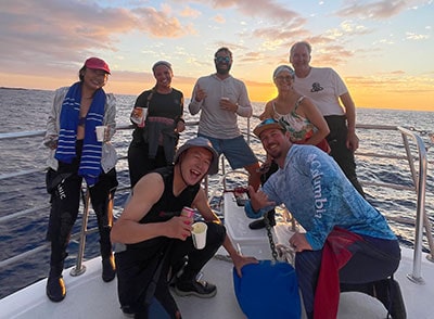 Group of divers and researchers celebrating coral reef conservation on a boat at sunset.