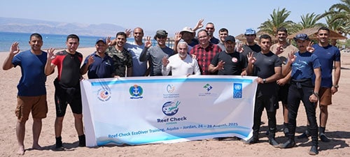 Diverse group of volunteers holding Reef Check banner on beach during coral reef conservation event.